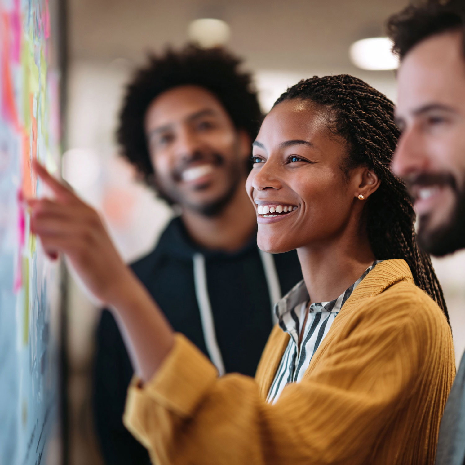 Une jeune femme et ses collègues travaillent sur un tableau rempli de post-it en souriant.